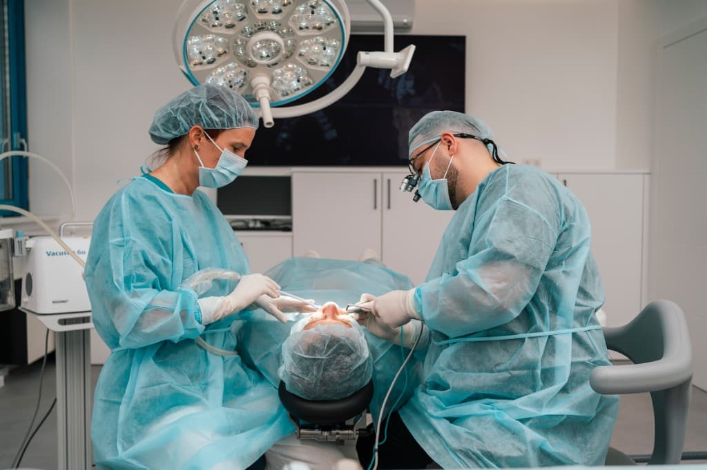 Zwei Behandelnde in steriler OP-Kleidung bei einer zahnärztlichen Behandlung unter OP-Licht in der Ordination Zahnwohl Penzing