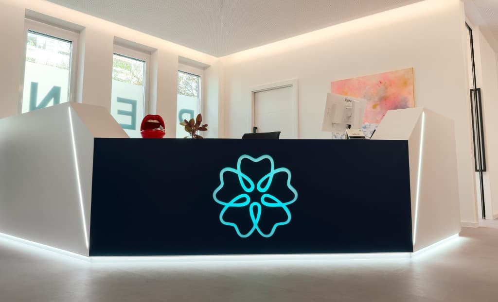 Empfang der Ordination Zahnwohl Penzing: moderne Rezeption mit leuchtendem Zahn-Logo in Türkis, dunkle Front, weiße Seitenelemente mit Lichtlinien, helle Räume, Wien 14
