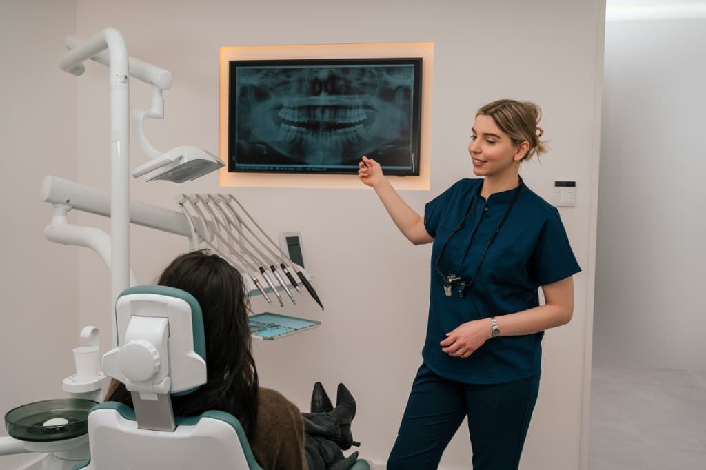 Zahnärztin erklärt einer Patientin am Monitor ein Panorama-Röntgenbild, helle Behandlungskabine mit Petrol-Stuhl und Instrumenten